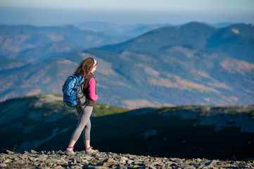 Naklejka premium Young woman tourist is climbing down on mountain plato with big backpack on. Beautiful mountains on background. Sunny autumn day.
