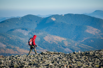 Male tourist walking on the rocky mountain ridge with beautiful mountains on background. Man is wearing red jacket and has trekking sticks and backpack on. Sunny fall day.