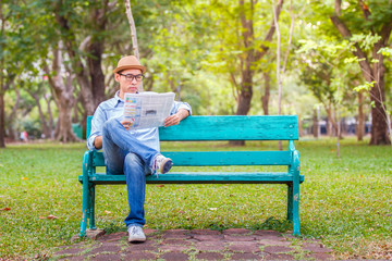 Asian young Man with hat sitting on a wooden bench and reading a newspaper in a park