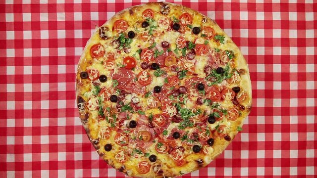 Rotating pizza with cheese, tomatoes and olives on the red tablecloth

