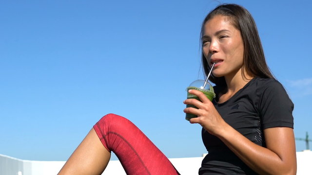 Happy sporty woman drinking healthy green vegetable juice. Young female is in sportswear. Beautiful fit lady relaxing after fitness workout training outdoors under blue sky.