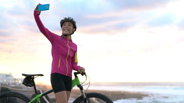 Happy young female cyclist taking selfie using smart phone while MTB mountain biking. Beautiful woman in sportswear is posing with bicycle. She is photographing using smart phone in nature with bike.