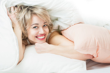 Obraz na płótnie Canvas Blonde woman lying in bed on a bed under a blanket. Wide beautiful smile with teeth. Charming relaxed girl