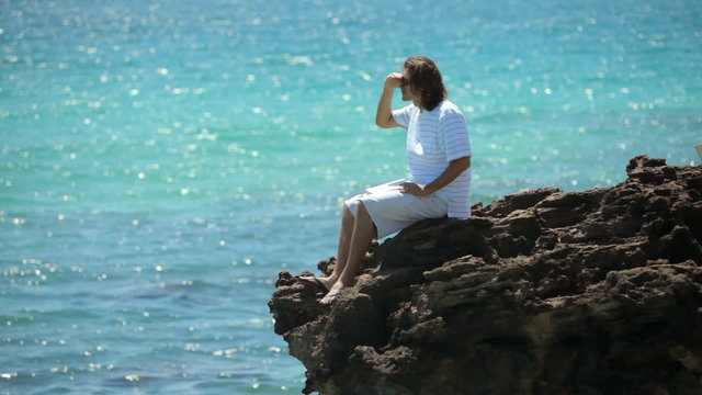 Man sits above ocean on rocky cliff with laptop computer