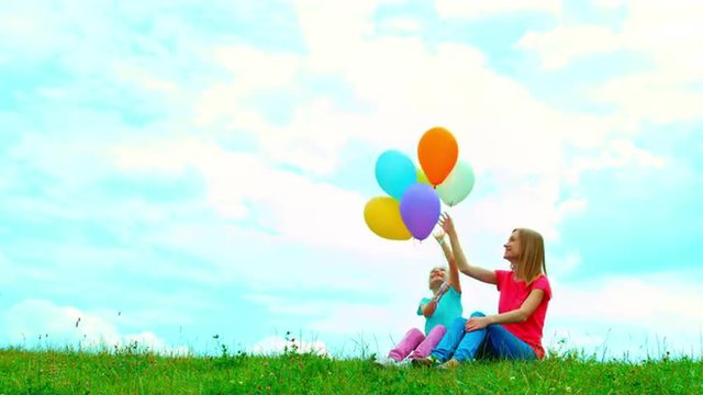 Mother and daughter with balloons sitting on the grass against the sky and playing with balloons