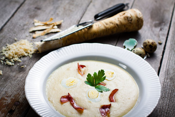 Creamy Horseradish soup with quail egg, black forest ham, decorated with leaf parsley. Soup in a deep plate on a gray wooden table in the background of grated horseradish root.