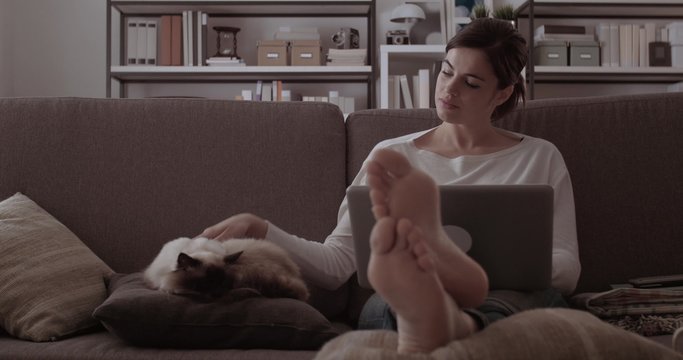 Young woman at home sitting on the sofa, she is working with a laptop and cuddling her beautiful birman cat sleeping next to her