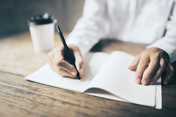 Man writing in blank diary and paper coffee cup on wooden table