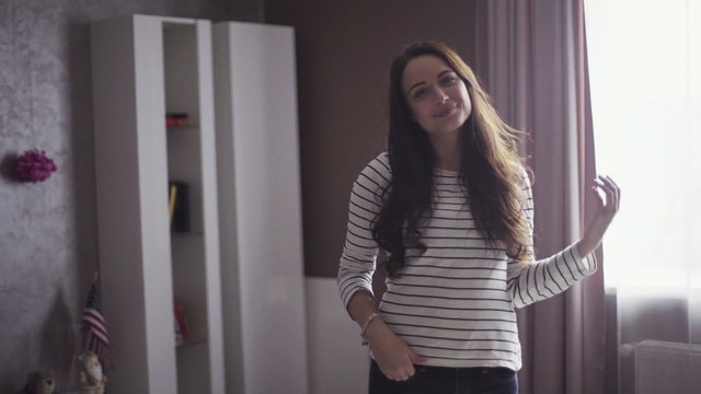 Slow motion portrait of a young cheeful woman who stands in a living room and touches her hair. 