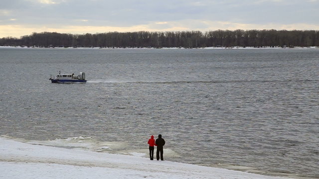 A man and a woman standing on the shore watching floating passenger hovercraft in the winter
