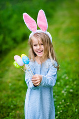 Cute laughing blond little girl wearing white and punk rabbit or bunny ears and blue knitted dress and holding bunch of painted colour eggs on sunny day in spring park. Easter celebrations