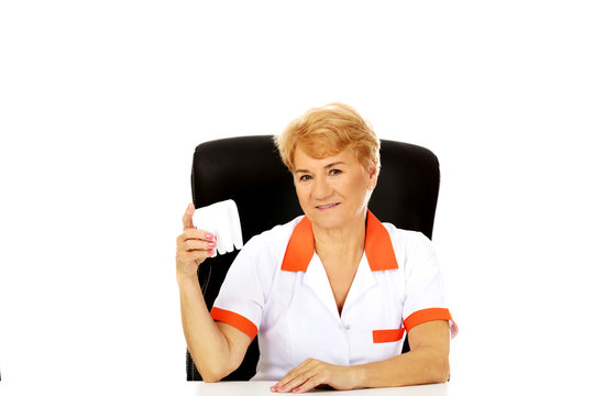 Smile elderly female dentist sitting behind the desk and holds big tooth model