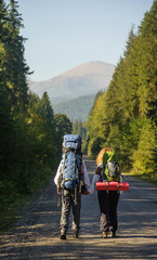 Obraz premium Rear view of couple backpackers walking on mountain road with backpacks, mountain on the background. Summer time.