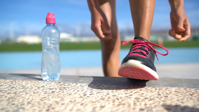 Sports woman runner getting ready for run tying laces of running shoes next to water bottle on athletic track and field background. Female athlete preparing for fitness cardio workout. Feet closeup.