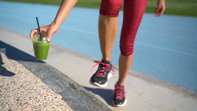 Fitness woman runner tying running shoes next to green smoothie cup on athletic track and field background. Female athlete getting ready for cardio workout run. Feet and legs closeup.