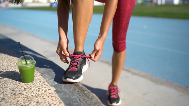 Athlete runner getting ready for run with green smoothie tying running shoes on outdoor athletic track and field with runners in the background. Legs and feet closeup.