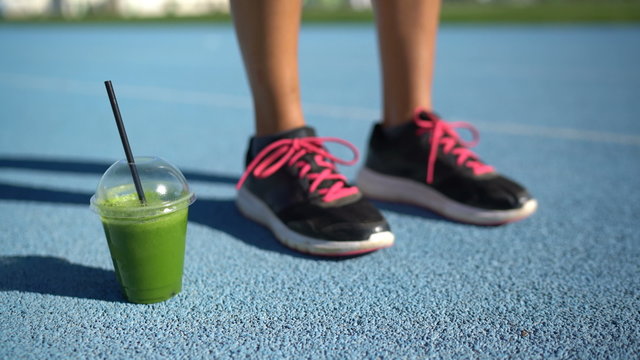 Fitness shoes and green smoothie juice cup on running track. Woman runner's feet ready for race with breakfast glass of organic vegetable juices for healthy cleanse or detox vegetarian diet.