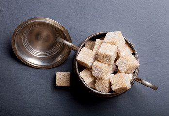 Old metal sugar bowl with pieces of brown sugar on black slate t