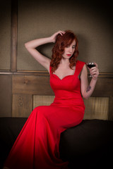 Elegant young redhead woman in a red dress, having a glass of red wine