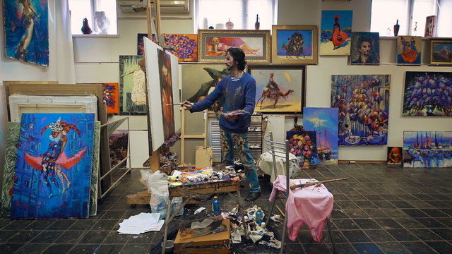 The artist paints with oil painting. In the studio, among his own works. 