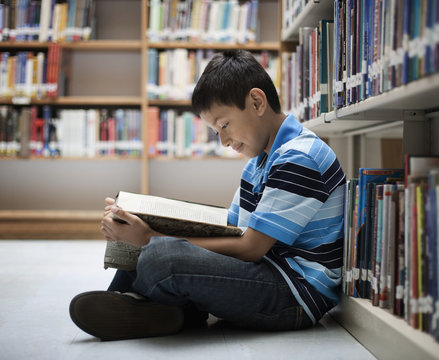 A boy sitting on the floor in a library reading a book,