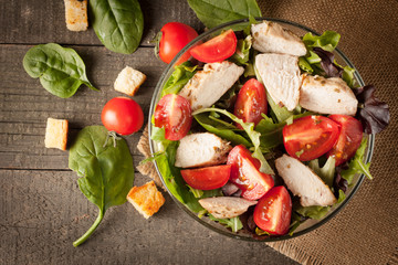 Fresh salad made of tomato, ruccola, chicken breast, arugula, crackers and spices. Salad in a white, transparent bowl on wooden background
