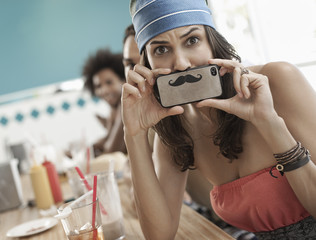 A woman holding a picture of a moustache on her smart phone just under her nose, 