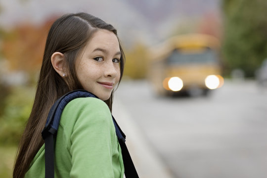 A young girl on the sidewalk, and a yellow school bus with headlights approaching, 