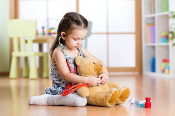 Little child girl with stethoscope and teddy bear sitting on floor, on home interior background