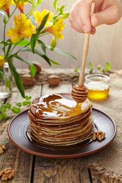 Spreading honey on stack of traditional homemade pancakes with nuts in clay dish on vintage wooden table background