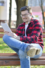 Obraz premium Portrait of young man looking at the camera and holding tablet PC. Happy man in casual working in the park while sitting on the bench.