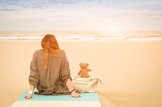 Young single woman sitting at beach with teddy bear looking at the sea - Solitude and loneliness concept with imaginary friendship and melancholic feelings - Warm vintage filter with enhanced sunshine