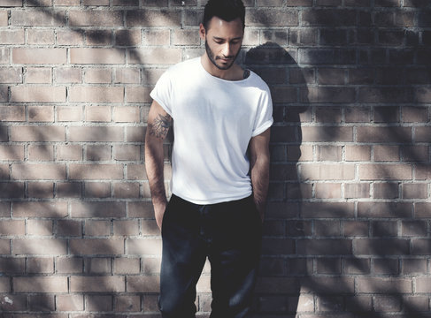Bearded man with tattoo wearing blank white tshirt and black jeans. Bricks wall background. Horizontal mockup, soft shadows