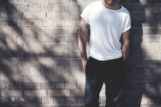 Photo bearded man with tattoo wearing blank white tshirt and black jeans. Bricks wall background. Wide mockup
