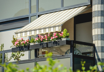 Balcony with awning opened and beautiful flowers - covered by sun-shield on a warm summer day - obrazy, fototapety, plakaty