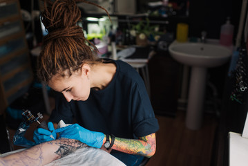 Fototapeta na wymiar Tattoo girl with dreadlocks on his head in the stuffing sleeve tattoo in black and white. Master works in a professional salon on a white mat and in sterile blue gloves.