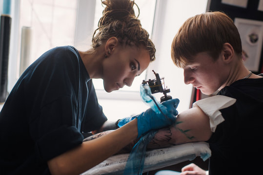 Tattoo girl with dreadlocks on his head in the stuffing sleeve tattoo in black and white. Master works in a professional salon on a white mat and in sterile blue gloves.