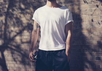 Naklejka na ściany i meble Photo modern man with tattoo wearing blank white tshirt. Stands in front of a brick wall. City street background. Horizontal 