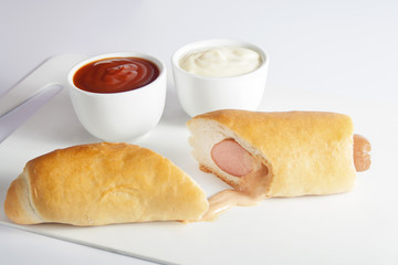 Hot dog cut in half,  placed on cutting board beside two small bowls with mayonnaise and ketchup.