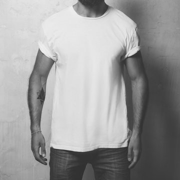 Closeup photo bearded angry man with tattoo wearing white blank t-shirt and black jeans, standing opposite concrete empty wall. Vertical ,mockup