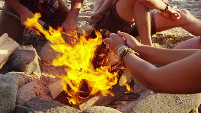 Young people at beach warm hands with campfire
