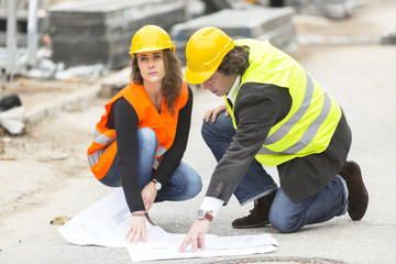 Male and female crouching architects with safety jackets and hardhats at construction site...