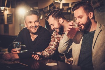 Cheerful old friends having fun with smartphone and drinking draft beer at bar counter in pub. - obrazy, fototapety, plakaty