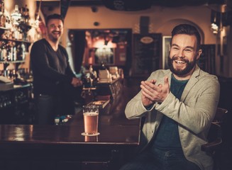 Cheerful stylish man having fun watching a football game on TV and drinking draft beer at bar counter in pub.