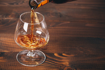 Pouring brandy or cognac from the bottle into the glass against wooden background