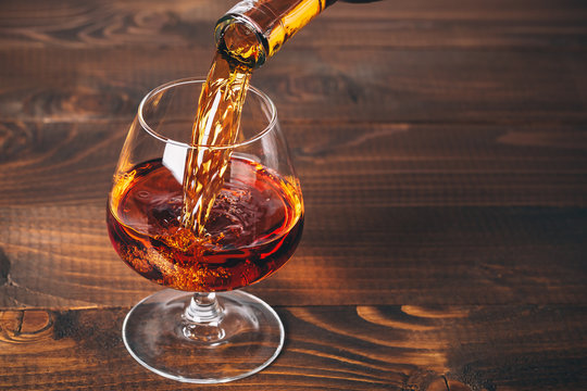 Pouring brandy or cognac from the bottle into the glass against wooden background