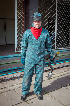 Adult man - paintball player standing equipped for the game in protective camouflage uniform and red scarf with marker gun