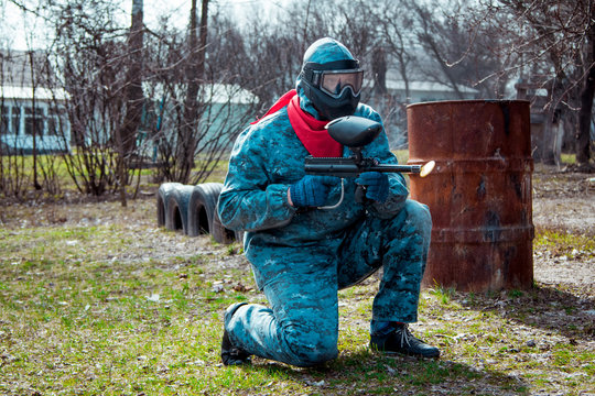 Adult man - paintball player posing equipped for the game in protective camouflage uniform and red scarf with marker gun