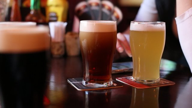 The waiter in bar brings and puts on table different grades of beers. On the background girl looking at her phone