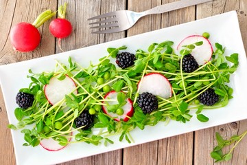 Healthy salad with pea shoots, radishes, blackberries on white rectangular plate on rustic wood...